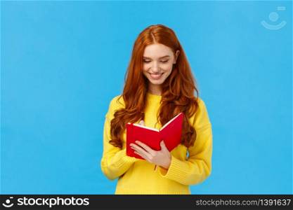 Waist-up portrait redhead female student write down thoughts in red lovely notebook, prepare grocery list, make schedule for college lectures, smiling as sharing thoughts in diary, blue background.. Waist-up portrait redhead female student write down thoughts in red lovely notebook, prepare grocery list, make schedule for college lectures, smiling as sharing thoughts in diary, blue background