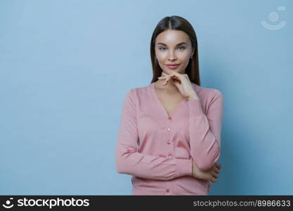 Waist up shot of beautiful young European woman keeps hand under chin, looks directly at camera, applies makeup, listens interlocutor carefully, wears casual jumper, isolated on blue background.