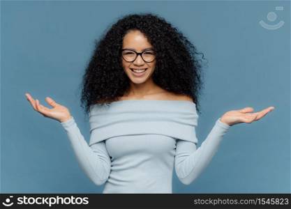 Waist up shot of happy young crisp Afro American woman raises both hands, pretends holding something, wears blue sweater, has naked shoulders, optical glasses, isolated over blue background.