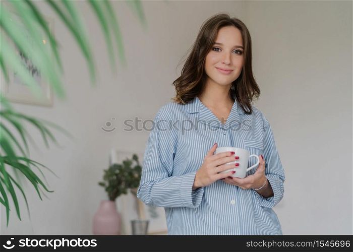 Waist up shot of pleasant looking young woman dressed in striped pyjamas, drinks hot beverage after awakening, has sparre time at home, stands indoor against cozy interior. People and rest concept