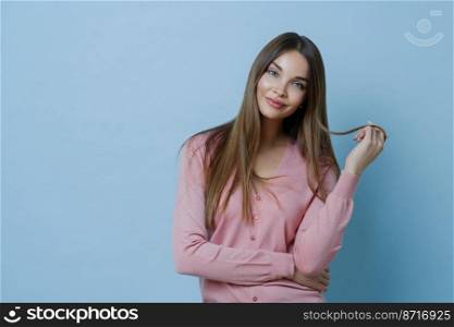 Waist up shot of pleasant looking young woman has beautiful long hair, tilts head, dressed in casual jumper, has perfect clean skin, advertises sh&oo for hair care, isolated on blue background.