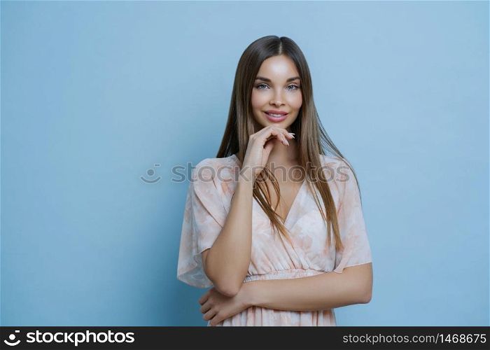 Waist up shot of pretty brunette European woman wears stylish summer dress, keeps hands partly crossed, holds chin, talks to coworker, isolated over blue background, cannot wait summer holidays