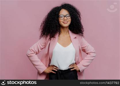Waist up shot of satisfied dark skinned female office worker keeps hands on her slim figure, has happy look, wears transparent glasses, poses over purple background, ready for talk with colleague