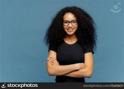 Waist up shot of smiling Afro American woman has arms folded, wears spectacles and casual black t shirt, enjoys nice conversation with interlocutor, poses over blue studio wall with blank space