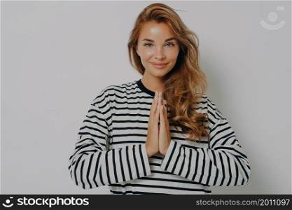 Waist up shot of smiling positive woman with folded hands in namaste gesture, pleased young female feeling love and gratefulness, making wish while posing isolated on grey studio wall background. Smiling positive young woman with folded hands in namaste gesture and smiling at camera