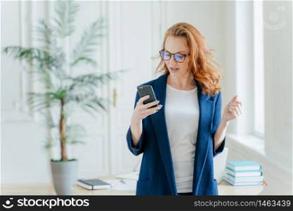 Waist up shot of successful businesswoman uses application on cell phone, reads recieved email from boss or colleague, dressed in formal clothes, poses over office interior, finds out financial news