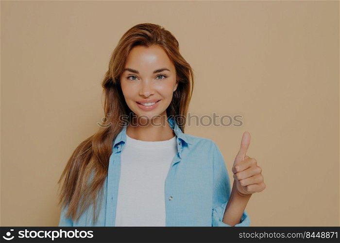 Waist up shot of supportive woman shows thumb up, cheers up her best friend, encourages for excellent efforts, wears blue oversized shirt and white tshirt, stands against beige background. Waist up shot of satisfied supportive woman shows thumb up