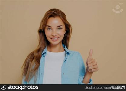 Waist up shot of supportive woman shows thumb up, cheers up her best friend, encourages for excellent efforts, wears blue oversized shirt and white tshirt, stands against beige background. Waist up shot of satisfied supportive woman shows thumb up