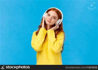 Waist-up shot tender and cute, lovely redhread woman in yellow sweater, tilt head, wear headphones, touching earphones as press to ears, listen music, smiling camera delighted, blue background.. Waist-up shot tender and cute, lovely redhread woman in yellow sweater, tilt head, wear headphones, touching earphones as press to ears, listen music, smiling camera delighted, blue background
