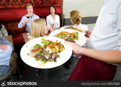 Waiter, carrying serveral dishes with food to the dinner guests waiting at the tables in a crowded restaurant