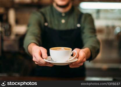 Waiter in black apron stretches a cup of coffee with cream in hands.