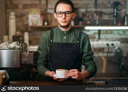 Waiter in black apron stretches a cup of coffee with cream in hands. Waiter in black apron stretches a cup of coffee