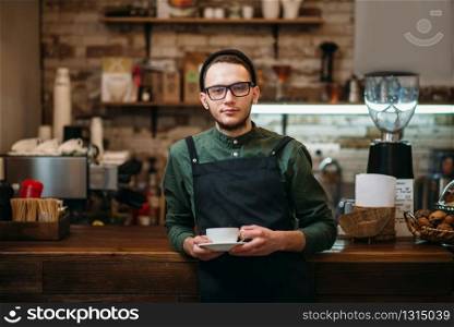Waiter in eyeglasses with a cup of coffee in hands has leaned the elbows on a bar counter.