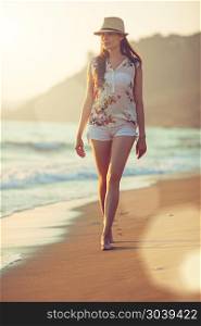 Walking barefoot on the beach. Young beautiful woman walking barefoot along the surf on the sand beach at sunset; lens flares