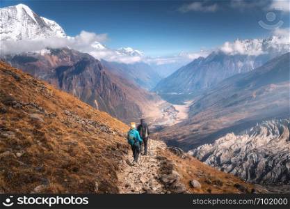 Walking people on the mountain trail at sunny day. Landscape with man and woman with backpacks, mountain valley, rocks with snowy peak in clouds, blue sky. Active people. Travel in Nepal. Trekking