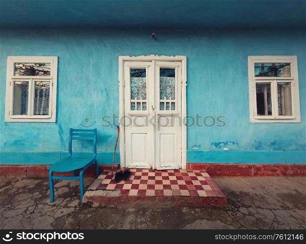 Walking stick, and a pair of old shoes and a chair on the doorstep of an aged house. Traditional rural building facade. Blue lime painted walls, white colored door and windows. Vintage home details.