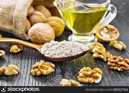 Walnut flour in a spoon, nuts on a table and in bag, oil in a glass sauceboat on background of dark wooden board