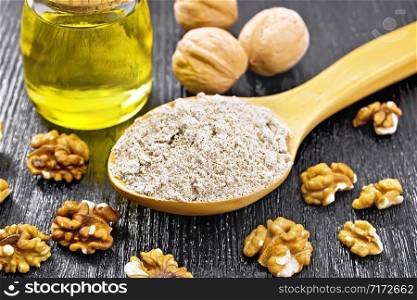 Walnut flour in a spoon, nuts on the table and oil in a glass jar on background of dark wooden board