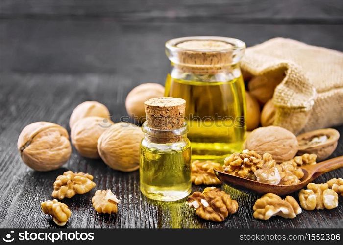 Walnut oil in a glass bottle and a jar, nuts in bag, spoon and on table against dark wooden board