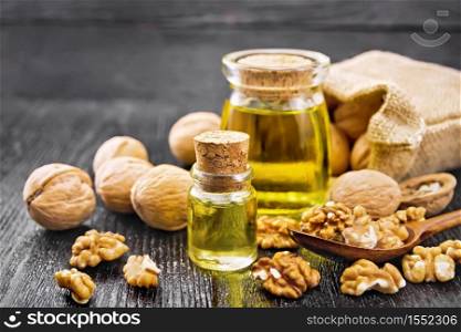 Walnut oil in a glass bottle and a jar, nuts in bag, spoon and on table against dark wooden board