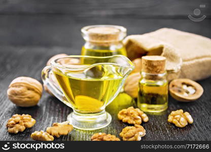 Walnut oil in a glass gravy boat and two banks, nuts in bag and on table on dark wooden board background