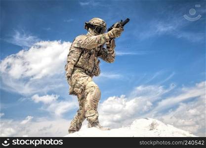 War game player in military camouflage uniform, equipped with tactical ammunition, aiming with service rifle replica while moving in sand with cloudy sky on background, during airsoft game event. Equipped airsoft player aiming with service rifle