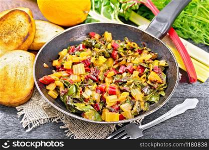 Warm chard salad with orange and onion in a frying pan on sackcloth, bread, fork on a black wooden board background