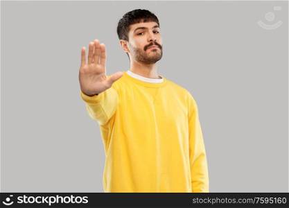 warning and people concept - serious young man in yellow sweatshirt showing stop gesture over grey background. serious young man showing stop gesture