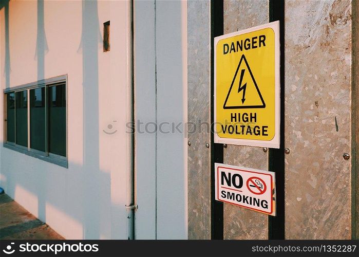 Warning danger high voltage sign and no smoking label on steel pipe cover with sunlight and electric pole's shadow on surface of white wall, sign and symbol with safety concept