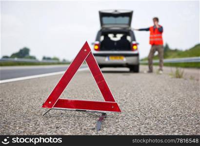 Warning tiangle behind a broken down car with a motorist calling for assistance. Focus on the triangle