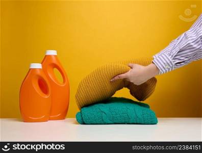 washed folded clothes and plastic orange large bottles with liquid detergent stand on a white table, yellow background. Routine homework