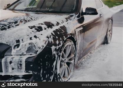 Washing off white soap foam with water from luxury black car at self-service car wash station outdoors, using high pressure washer and detergent for professional vehicle cleaning. Washing off white soap foam from car with high pressure washer outdoors