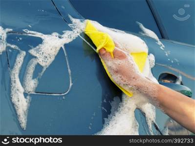 Washing the gray car with a yellow rag in soapy foam. A woman's hand wipes the side of the car. Washing the gray car with yellow rag in soapy foam