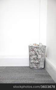 Wastebasket full of crumpled paper in corner against white wall