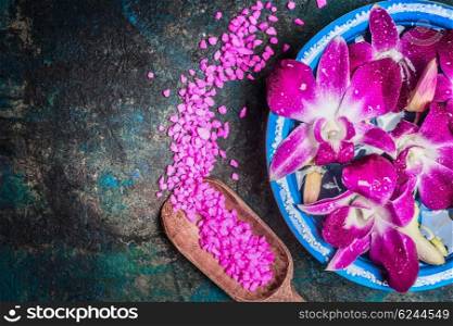 Water bowl with pink orchid flowers on dark background with shovel of sea salt, top view, place for text. Spa, wellness or body care concept.