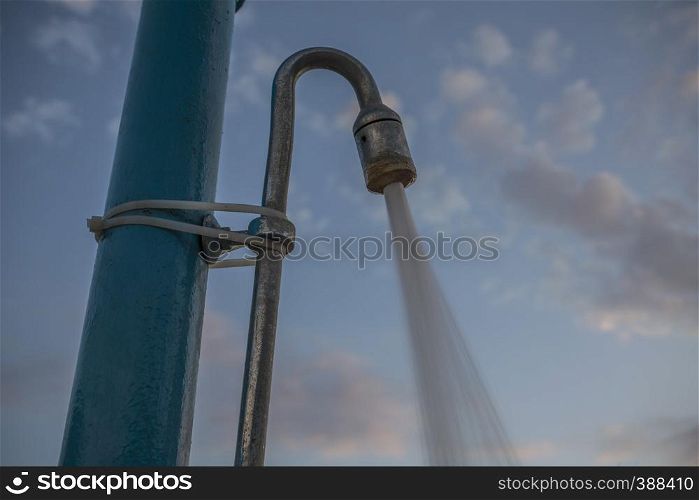 Water Pouring from Shower Head. Public Shower for Outdoor swimming under the Blue Cloudy Sky. Closeup Angle Shot of Pipe Tied to Pole by Plastic Strip Seal. Water Pouring from Shower Head. Public Shower for Outdoor swimming under the Blue Cloudy Sky. Closeup Angle Shot of Pipe Tied to Pole by Plastic Strip Seal.