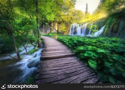Waterfall and wooden path in green forest in Plitvice Lakes, Croatia at sunset in summer. Colorful landscape with trail in blooming park, trees, water lilies, river, sunbeams in spring. Trail in woods