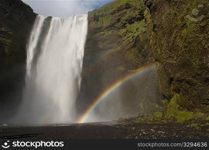 Waterfall cascading over a green rock cliff, with a double rainbow coming from the mist from the lake