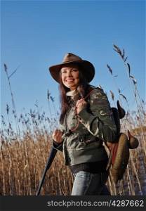 Waterfowl hunting, smiling female hunter carry a shotgun and a decoys, reeds and blue sky on background