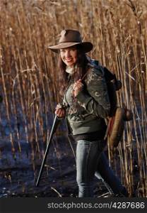 Waterfowl hunting, smiling female hunter carry a shotgun and a decoys, shore and reeds on background