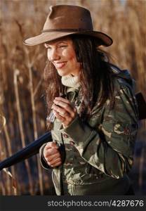 Waterfowl hunting, smiling female hunter carry a shotgun and she use a duck call, shore and reeds on background