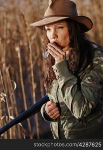 Waterfowl hunting, the female hunter carry a shotgun and she use a duck call, shore and reeds on background