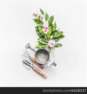 Watering can with gardening tools and green bunch of twigs with blossom decoration on white desk background, top view