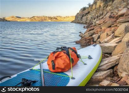 waterproof duffel on a deck of an inflatable stand up paddleboard, rocky shore of mountain lake - Horsetooth Reservoir in northern Colorado, traip and expedition concept