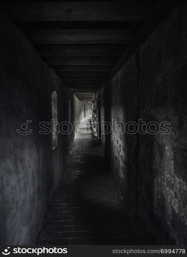 Weathered passageway in low light, in a medieval building, with beams of light. A long hallway in shadow with light at the end. Vintage architecture.