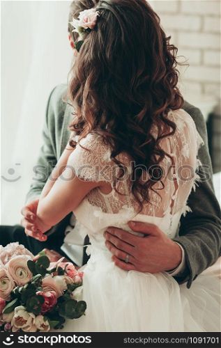 Wedding bouquet with blurred bride and groom in the background, holding each other in arms, sitting on the bed, view from bride back