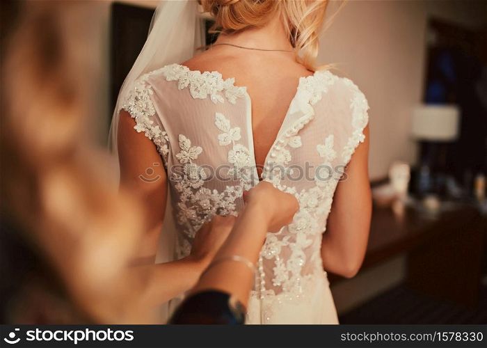 Wedding. Bridesmaid preparing bride for the wedding day. Bridesmaid helps fasten a dress before the ceremony. Luxury bridal dress close up. Wedding. Bridesmaid preparing bride for the wedding day. Bridesmaid helps fasten a dress before the ceremony. Luxury bridal dress close up.