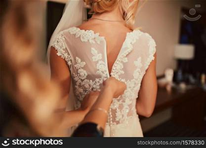 Wedding. Bridesmaid preparing bride for the wedding day. Bridesmaid helps fasten a dress before the ceremony. Luxury bridal dress close up. Wedding. Bridesmaid preparing bride for the wedding day. Bridesmaid helps fasten a dress before the ceremony. Luxury bridal dress close up.