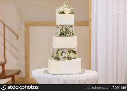 Wedding cake. Close-up photo of a beautiful white three-tiered wedding cake decorated by flowers and greenery.. Wedding cake. Close-up photo of a beautiful white three-tiered wedding cake decorated by flowers and greenery