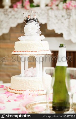 Wedding cake decorated with two grooms, on a reception table with champagne and rose petals.